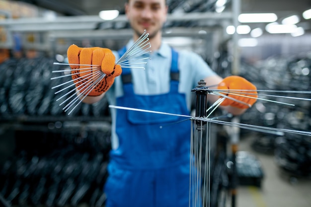 Worker in uniform shows new bicycle spokes on factory. Bike wheels assembly in workshop, cycle parts installation