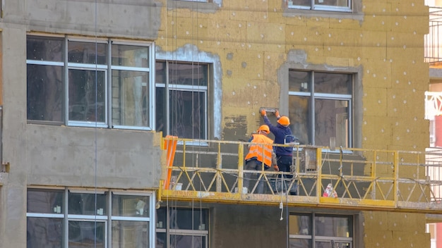 Workers are insulating the wall of a building construction site with working men on the elevator pan