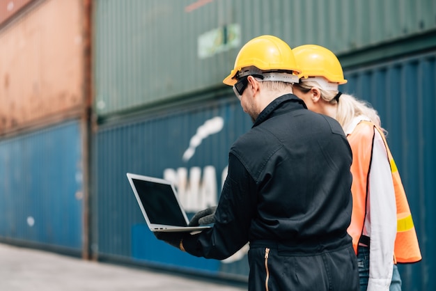 Workers teamwork man and woman in safety jumpsuit with yellow helmet using laptop and talking at cargo container.female and male work in dock warehouse. transport import,export logistic industrial