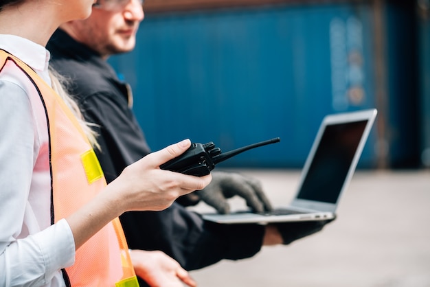 Workers teamwork man and woman in safety jumpsuit workwear with yellow hardhat and use laptop check container at cargo shipping warehouse.