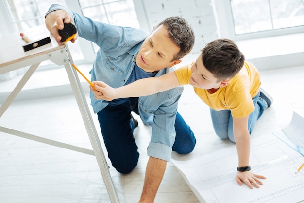 Working as team. Pleasant young father using a measuring tape and figuring out the length of a table leg together with his son while working on a blueprint