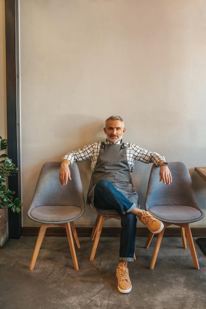 Working break. Calm middle aged man in apron resting sitting relaxed on chair looking confidently at camera in cafe