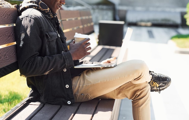 Working by using laptop Close up view of african american man in black jacket outdoors in the city sitting on bench