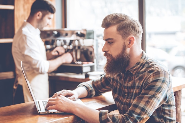 Working in cafe. Side view of young handsome bearded man using his laptop while sitting at bar counter at cafe with barista at the background