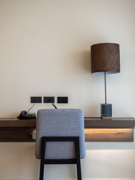 Working corner back view of empty grey cozy fabric chair seat black telephone desk lamp and sockets decorated on wood shelf on white wall background in hotel room with copy space vertical style