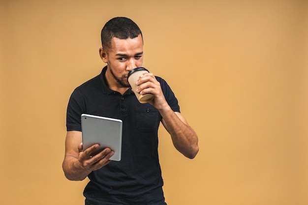 Working on digital tablet Portrait of happy smiling young African american black man holding digital tablet while standing isolated over beige background Drinking coffee