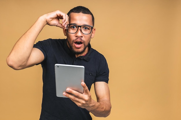 Working on digital tablet Portrait of shocked amazed surprised young African american black man holding digital tablet while standing isolated over beige background