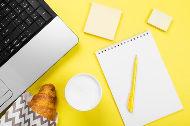 Working morning. Breakfast at work - cappuccino, croissant, notebook on yellow space. Top view