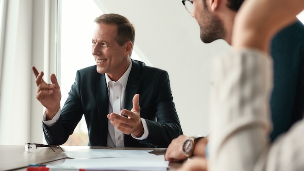 Working together. Handsome and happy mature man in formal wear gesturing and smiling while sitting with his colleagues in the office. Business people. Business meeting