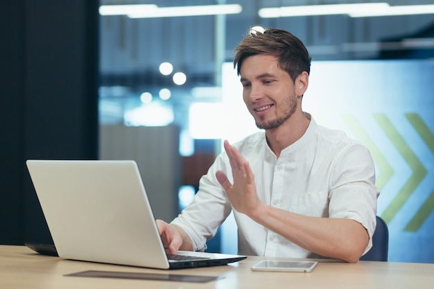 Working video call in the office Young male businessman freelancer sitting at a desk in the office on a laptop talking through the camera on a video call explaining something waving his hands