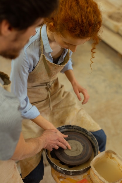 Photo working on wheel. curious ginger girl sitting on a chair and learning pottery wheel technique with assistant of dark-haired master