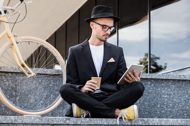 Working with technology out of office. Young man in stylish casual suit talking on the phone in urban area, sitting next to a commuter bike