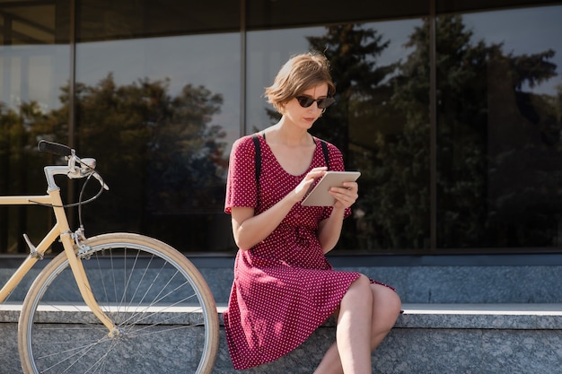 Working with technology out of office. Young woman in stylish retro dress  working with a tablet computer outdoors in urban area, sitting next to a commuter bike