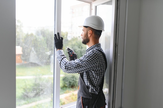 Photo workman in overalls installing or adjusting plastic windows in the living room at home