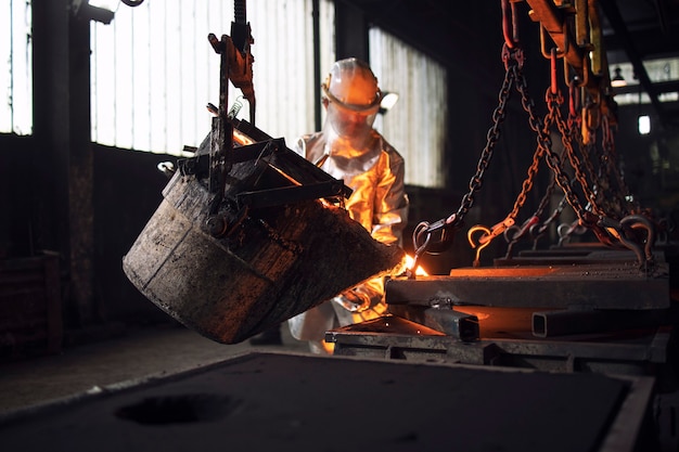 Workman in protective suit hardworking with liquid metal in foundry.