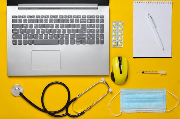 Workplace of a modern doctor. Laptop, wireless mouse, notebook, stethoscope, pills on a yellow pastel background. Top view, minimalist trend