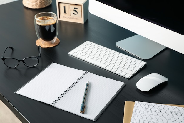 Workplace with computer, glass of coffee and empty notebook on wood, closeup