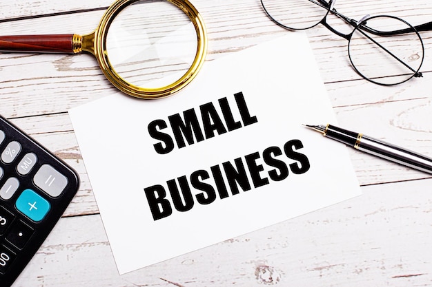 A workplace with a pen glasses a calculator a magnifying glass and a white sheet of paper with the text of the SMALL BUSINESS on a white wooden table Business concept