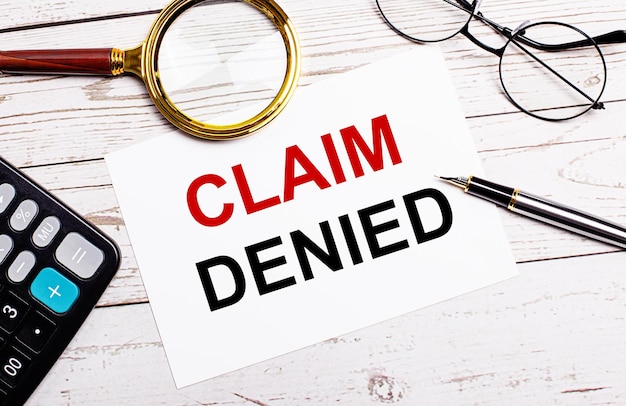 A workspace with a pen, glasses, a calculator, a magnifying glass and a white sheet of paper with the text of the CLAIM DENIED on a white wooden table. Business concept.