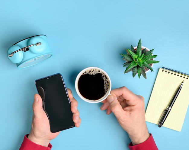 Workspace with smartphone and coffee in male hands on blue office desk table with copy space. Flat lay design.