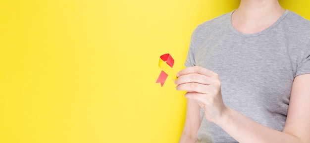 World Hepatitis Day concept. Girl in gray T-shirt holds in her hand awareness symbol red-yellow ribbon. Yellow background. Copy space