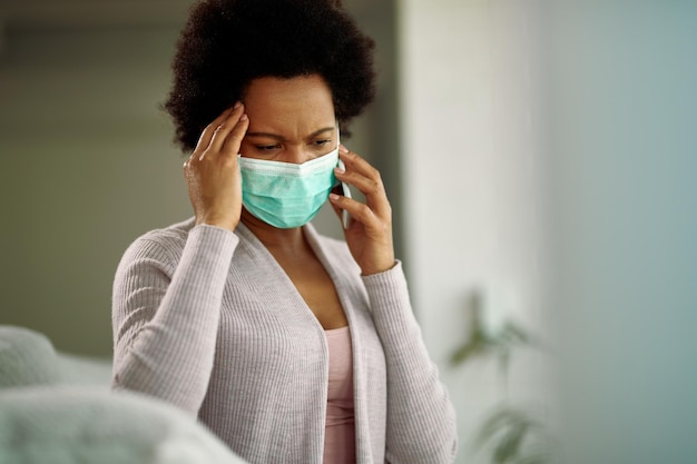 Worried African American woman wearing protective face mask while talking on mobile phone at home