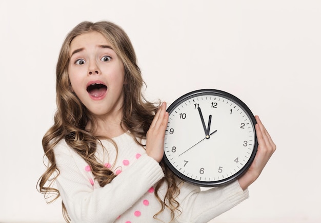 Worried little girl with clock at white studio background, copy space. Being late, time is the most precious resource concept