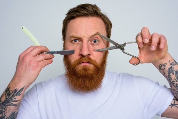 Worried man with scissors and blade is ready to cut the beard