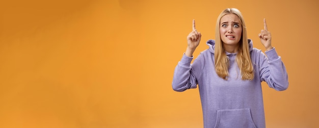 Photo worried perplexed unsure cute blond woman presenting project standing awkward afraid humiliation crining scared pointing up raised index finger waiting reply nervously orange background