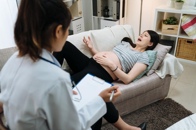 Worried pregnant girl lying on sofa next to asian female counselor during therapy in home living room. psychiatrist doctor examining to maternity woman patient. giving counseling and suggestion.