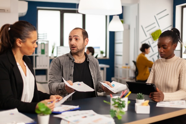 Worried startup diverse team discussing about financial results, discussing sitting at desk in office, planning next strategy holding tablet searching solutions