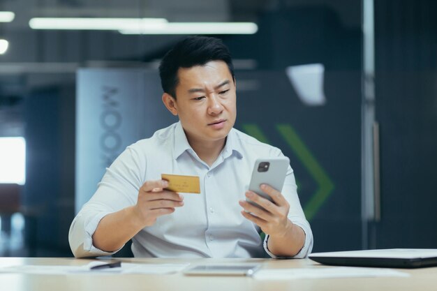 Worried young asian man sitting at desk in office and holding phone and blocked credit card