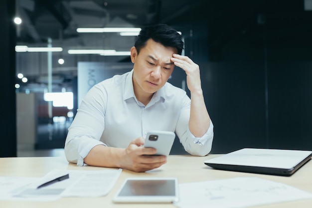 Worried young asian man sitting in office at desk and using mobile phone upset he looks at the