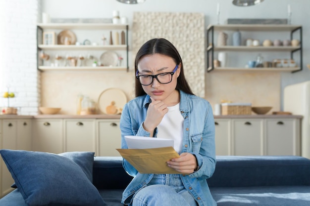 A worried young asian woman received a letter with bills for credit rent mortgage sitting at home on