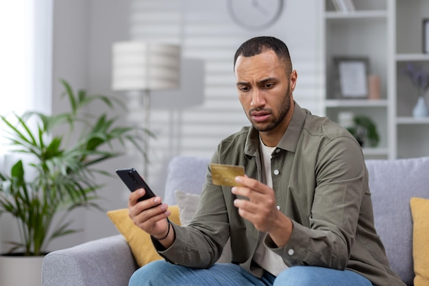 Worried young hispanic man sitting on sofa at home and looking at credit card upset holds the phone