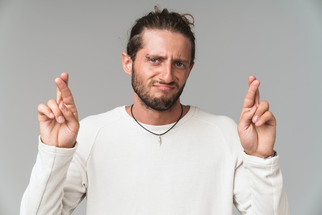 Worried young man standing isolated on gray, holding fingers crossed for good luck