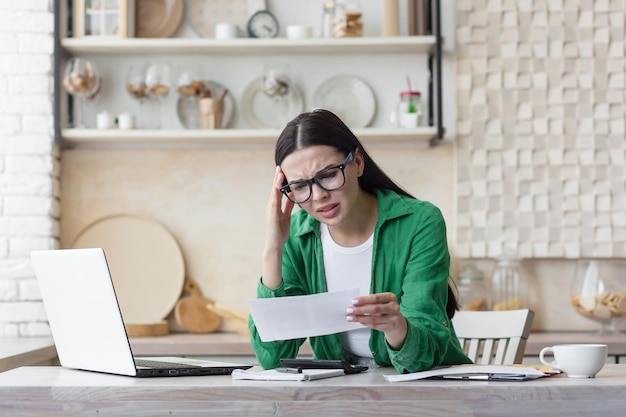 Worried young woman reading a received letter while sitting in the kitchen at home