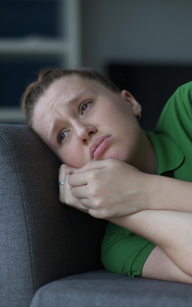 Worried young woman sit on sofa look aside lean forward with folded hands feel fear anxiety teen