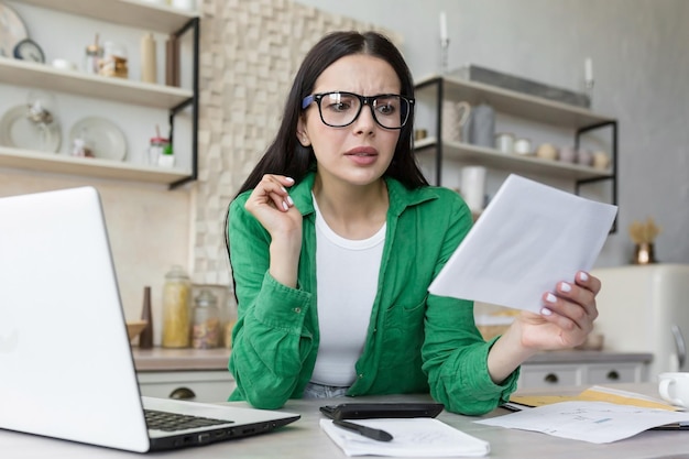 Worried young woman sitting in the kitchen at the table at home with a laptop holds a letter a