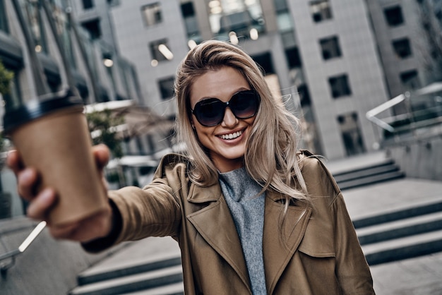 Would you like some coffee? Attractive young woman carrying disposable cup and looking at camera while standing outdoors