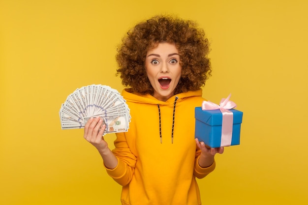 Wow cashback thrift shopping Portrait of surprised curlyhaired woman in urban style hoodie holding money dollar banknotes and gift box expressing amazement indoor studio shot yellow background