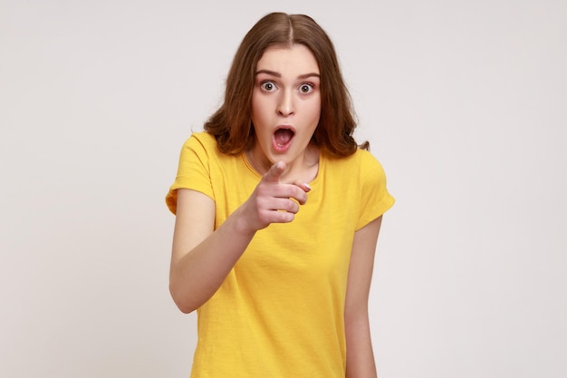 Wow hey you. Amazed beautiful brown haired female of young age in yellow T-shirt pointing to camera and looking surprised, keeping mouth open. Indoor studio shot isolated on gray background.