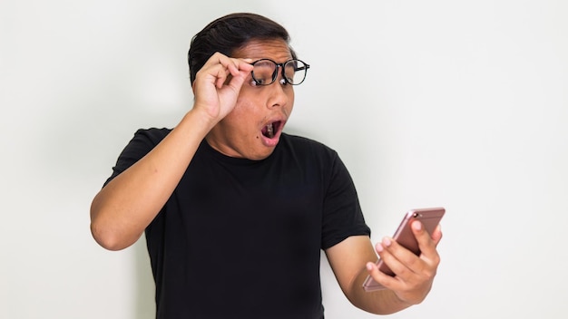 Wow and surprised expression of young Asian Malay man with spectacles in black t-shirt by looking something he saw on the mobile screen on isolated white background.