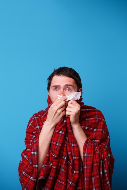 A wrapped in a blanket sick man is blowing his nose in white handkerchief