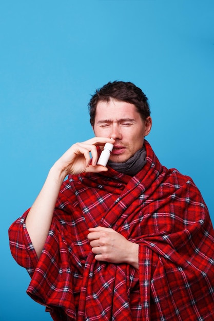 A wrapped in a blanket sick man is standing and using nasal spray