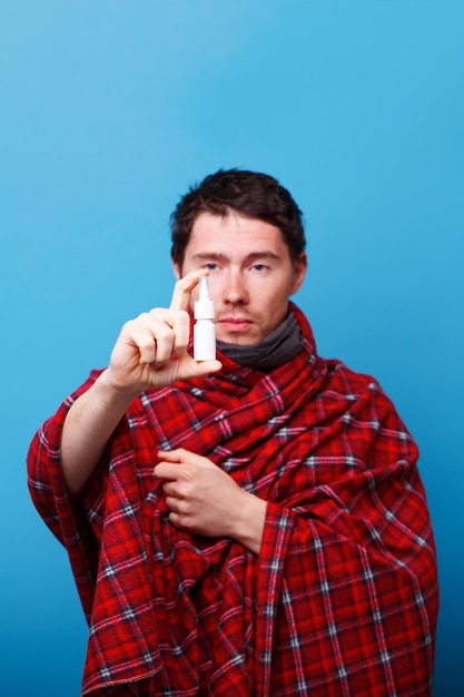 A wrapped in a blanket sick man is standing with nasal spray in his hand