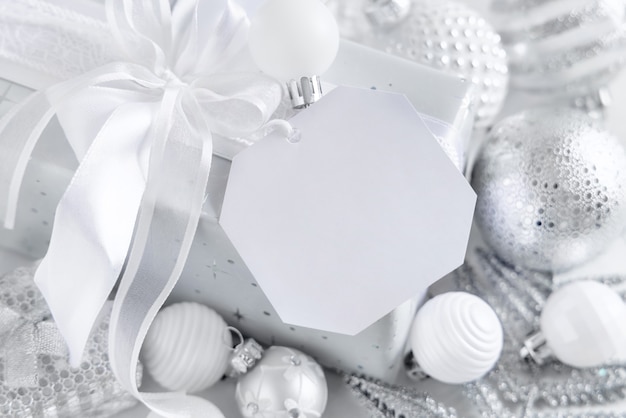 Wrapped present with a white bow and paper gift tag on a white table with white and silver Christmas decorations close up. Winter composition with blank label card, Mockup, copy space