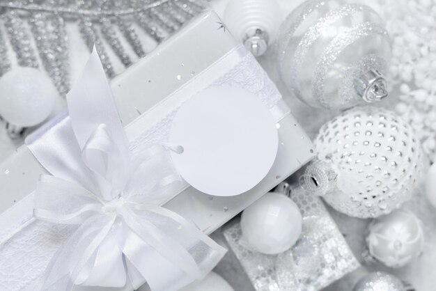 Wrapped present with a white bow and round paper gift tag on a white table with white and silver Christmas decorations top view. Winter composition with blank label card, Mockup, copy space