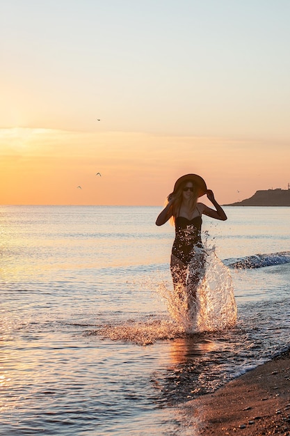 XAA happy girl in a swimsuit and a hat runs along the sea at dawn