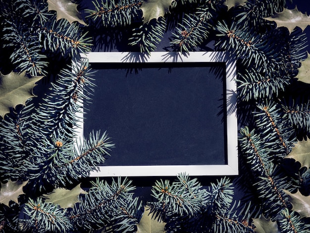 Xmas fir twigs and holly leaves natural undecorated frame around lackboard chalk board Christmas background in black and dark green